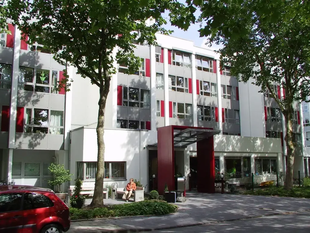 Ein modernes vierstöckiges Mehrfamilienhaus mit roten und weißen Platten, Bäumen davor und einer Person, die auf einer Bank in der Nähe des Eingangs sitzt, in der Nähe des Pflegezentrums Herten.