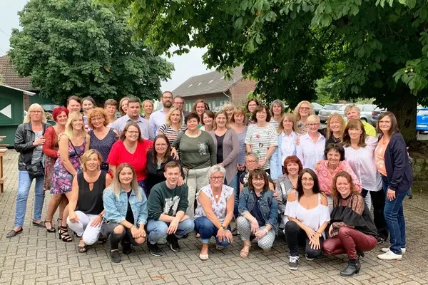 Eine Gruppe von etwa 40 Erwachsenen posiert gemeinsam im Freien auf einer gepflasterten Fläche unter Bäumen und lächelt in die Kamera vor dem Pflegezentrum Herten.