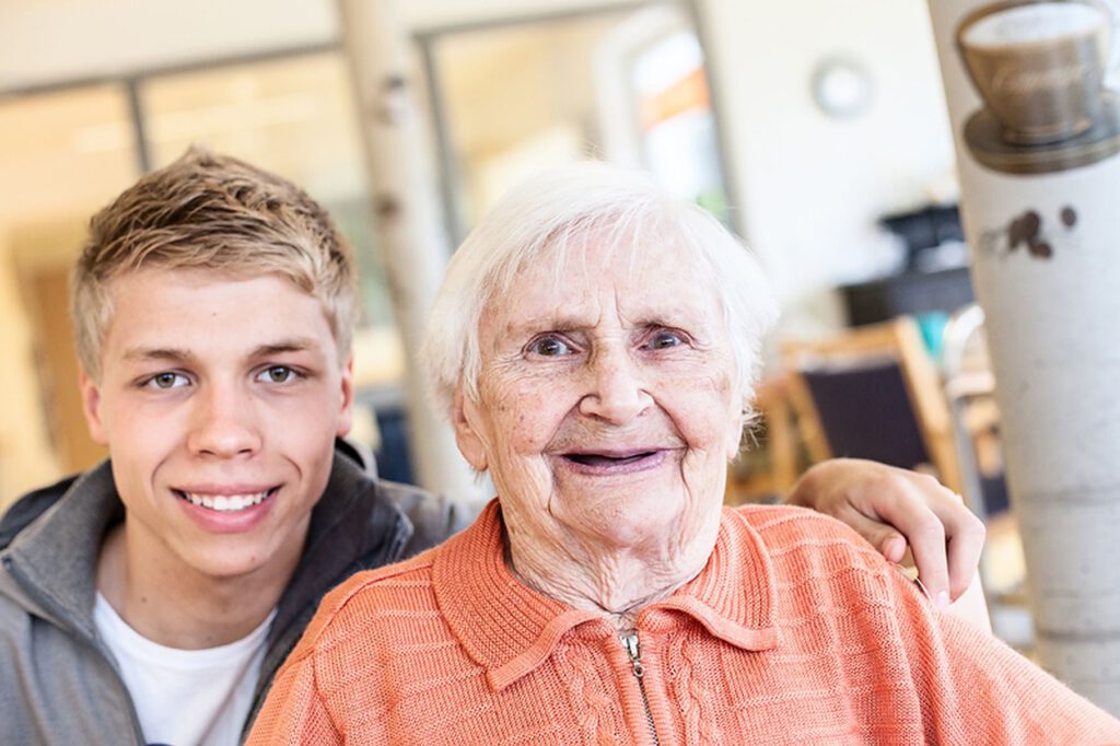 Ein junger Mann mit kurzen blonden Haaren sitzt neben einer älteren Frau mit weißen Haaren und einem orangefarbenen Pullover, die beide in die Kamera lächeln.