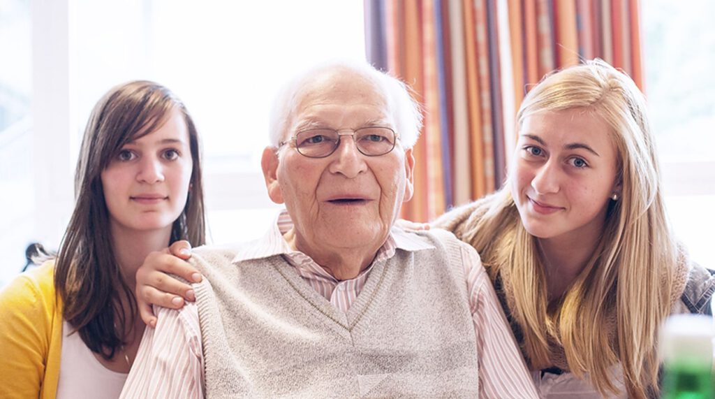 Ein älterer Mann mit grauem Haar und Brille sitzt zwischen zwei jungen Frauen, die der Kamera zugewandt sind, in einem Innenraum mit natürlichem Licht aus einem Fenster hinter ihnen.