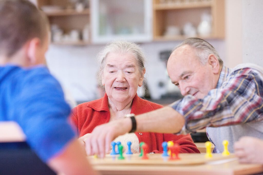Zwei ältere Erwachsene und eine jüngere Person sitzen an einem Tisch und spielen ein Brettspiel mit bunten Figuren in einer hellen, wohnlichen Umgebung.