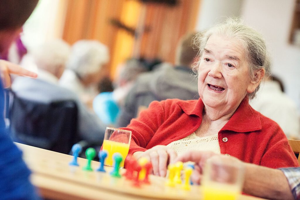 Eine ältere Frau in einer roten Strickjacke sitzt an einem Tisch, spielt ein Brettspiel mit bunten Figuren und hat zwei Gläser Orangensaft vor sich stehen.
