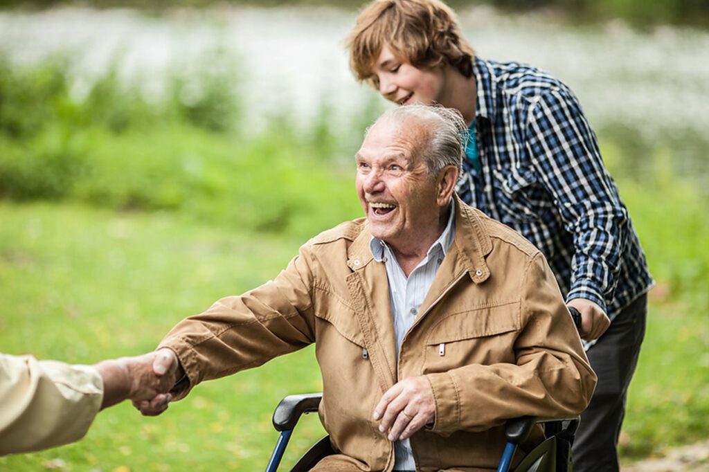 Ein älterer Mann im Rollstuhl, der lächelt und jemandem die Hand schüttelt, steht im Freien, während eine jüngere Person hinter ihm steht. Im Hintergrund sind Gras und Wasser zu sehen.