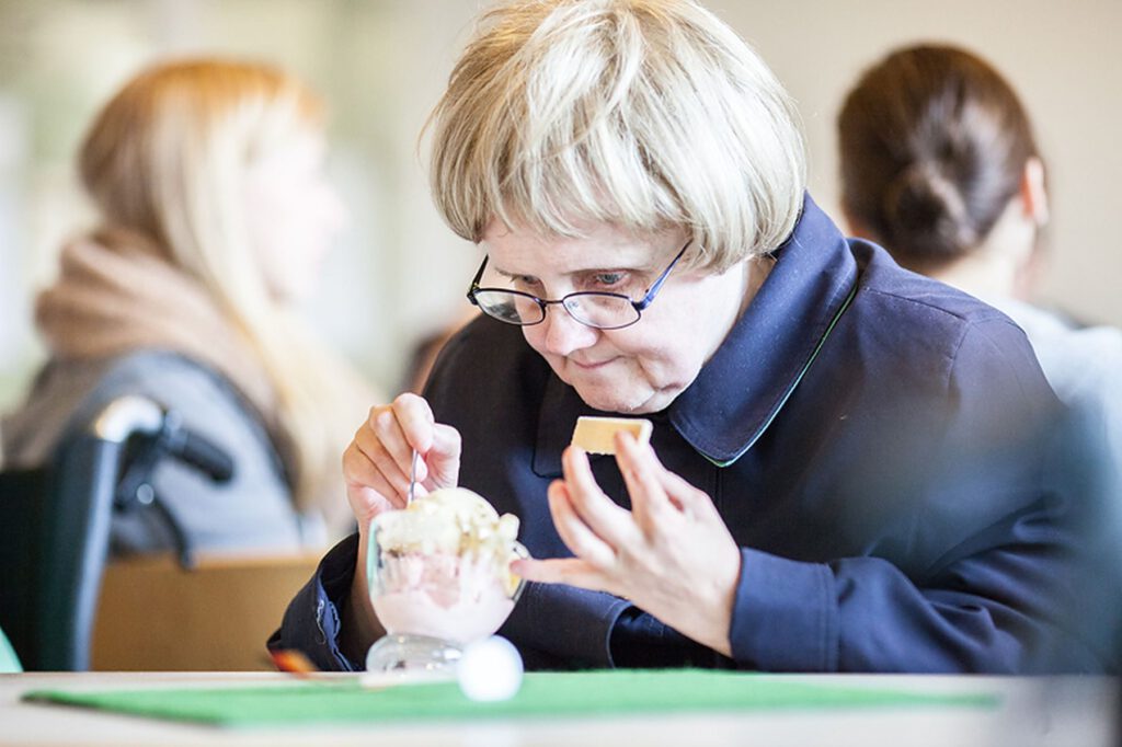 Eine ältere Frau mit Brille isst an einem Tisch Eis aus einer Glasschale, während andere Personen im Hintergrund verschwommen zu sehen sind.