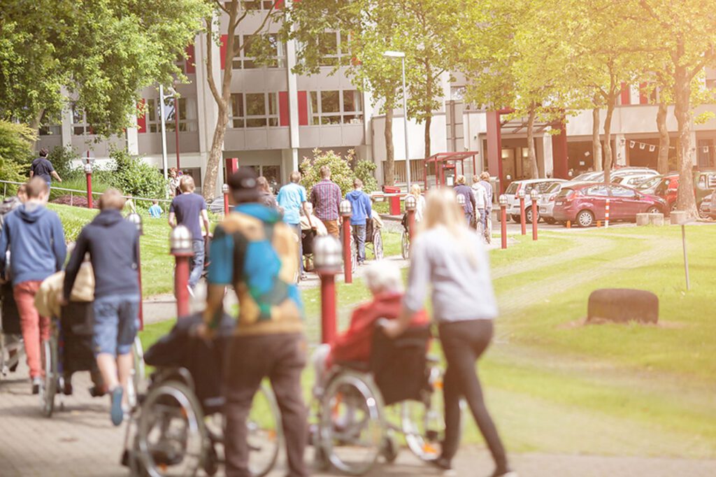 Menschen, einige im Rollstuhl, andere zu Fuß, bewegen sich an einem sonnigen Tag auf einem Weg vor einem Gebäude, umgeben von Bäumen und geparkten Autos.