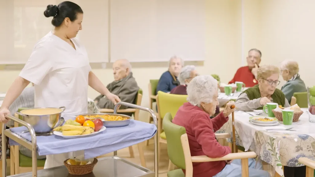 Eine Pflegekraft steht an einem Essenswagen mit Obst und Geschirr, während ältere Menschen an Tischen in einem Essbereich einer Pflegeeinrichtung sitzen.
