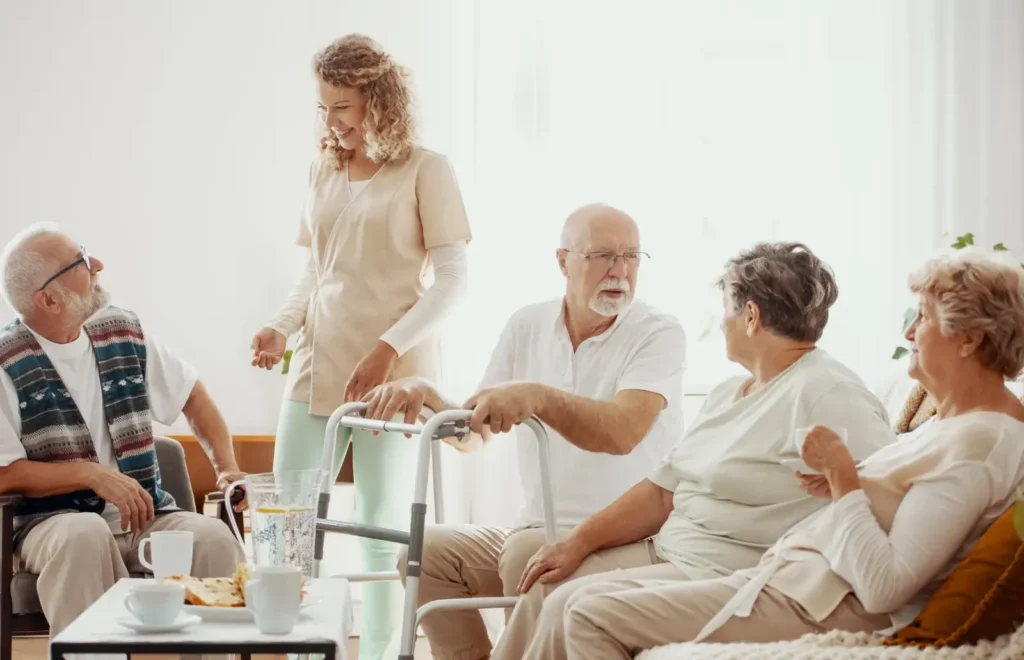 Eine Gruppe älterer Menschen sitzt in einem hellen Raum und unterhält sich mit einer Pflegerin, die in der Nähe steht. Ein Mann benutzt eine Gehhilfe. Auf dem Tisch vor ihnen stehen Getränke und Snacks.
