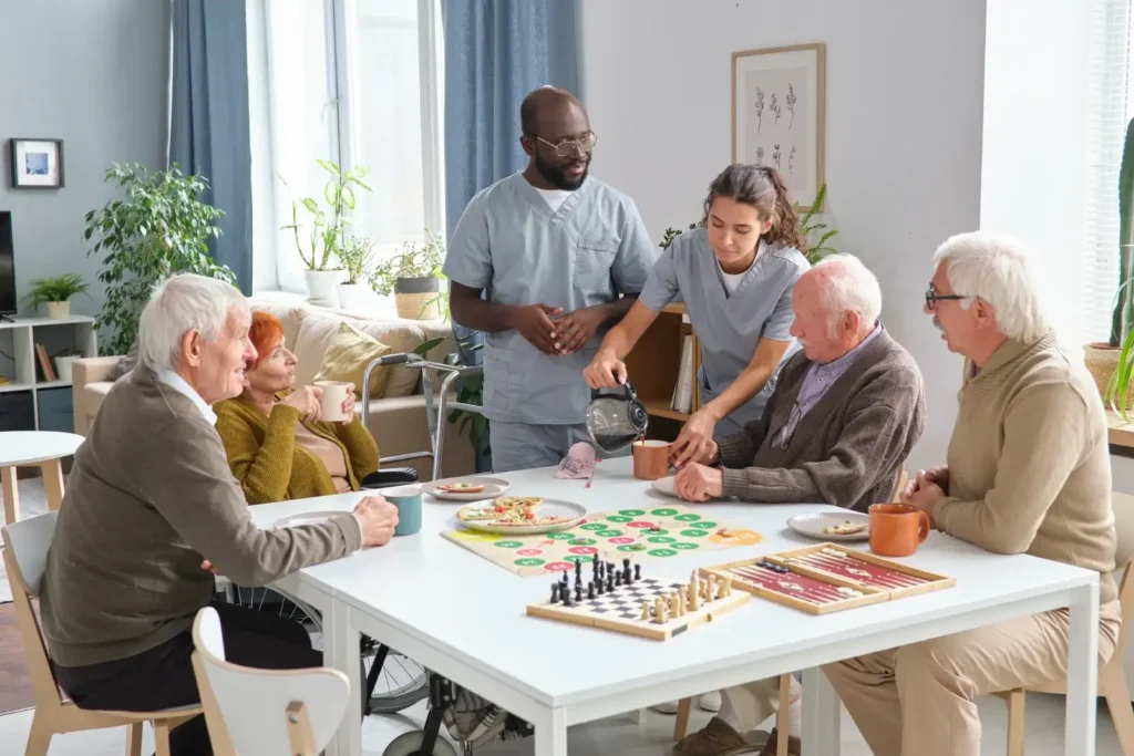 Zwei Betreuer helfen vier älteren Menschen, die in einem gut beleuchteten, gemütlichen Raum um einen Tisch mit Brettspielen, Snacks und Getränken sitzen.