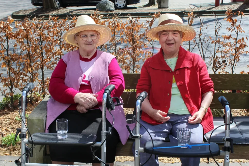Zwei ältere Frauen mit Strohhüten und bunten Pullovern sitzen auf einer Bank im Freien, jede mit einem Rollator und einem Glas Wasser vor sich.