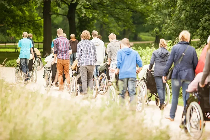 Eine Gruppe von Menschen geht draußen auf einem Weg spazieren, einige schieben Personen in Rollstühlen, umgeben von Bäumen und Grünflächen.
