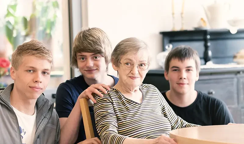 Eine ältere Frau sitzt an einem Tisch, dicht hinter ihr stehen drei junge Männer, die alle in einem hellen Innenraum in die Kamera schauen.